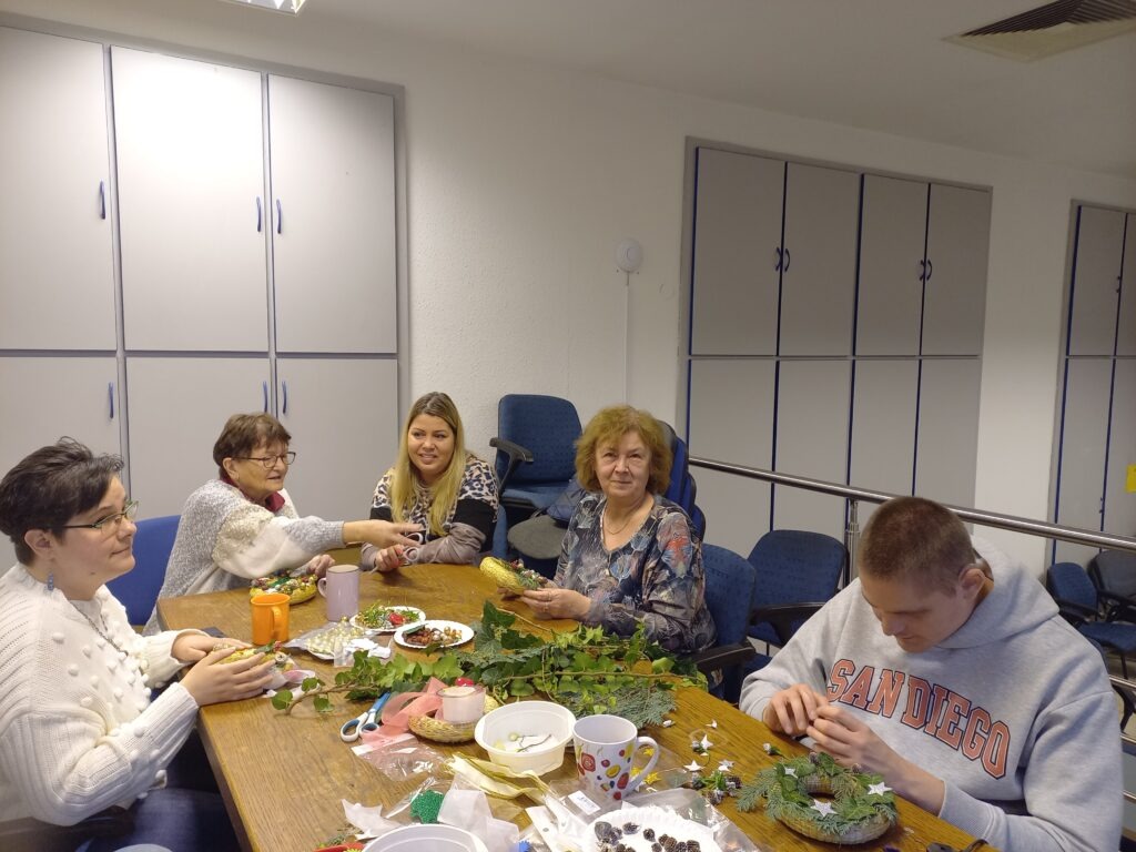 Asztal körül ülnek a résztvevők, előttük koszorúalap, fenyőág, toboz és más alapanyagok, díszítő elemek az alkotáshoz