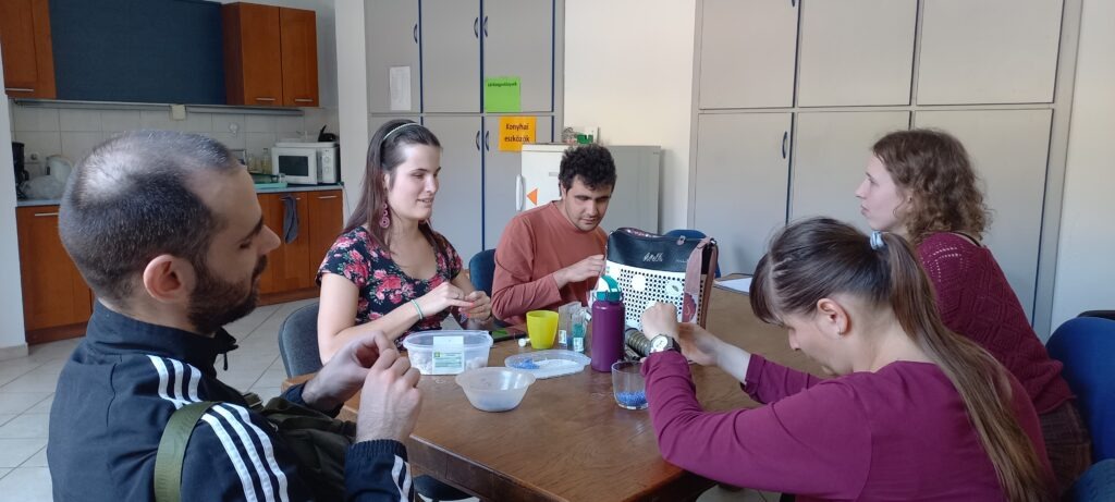 Az asztal körül ülnek a résztvevők, gyöngyöt fűznek, vidáman beszélgetnek, tákákban a lila gyöngyök, amiveldolgoznak