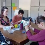 Az asztal körül ülnek a résztvevők, gyöngyöt fűznek, vidáman beszélgetnek, tákákban a lila gyöngyök, amiveldolgoznak