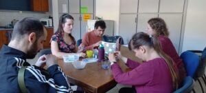 Az asztal körül ülnek a résztvevők, gyöngyöt fűznek, vidáman beszélgetnek, tákákban a lila gyöngyök, amiveldolgoznak