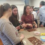 A résztvevő klubtagok egy márvány tetejű asztalt körbeállnak és csokoládét készítenek