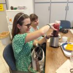 Elsőként próbálja ki a Smoothie készítő gépet a klub főszervezője, profin illeszti a turmixgépre az üveget, mellette egy másik résztvevő ül