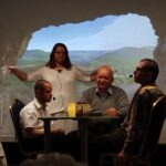 Péter és Pál az asztalnál ülnek, mögöttük a csend szétterített fátylával, háttérben szépen látszik a kivetített táj