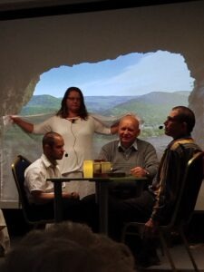 A színi előadás egyik jelenete: Péter és Pál az asztalnál ülnek, mögöttük a csend szétterített fátylával, háttérben szépen látszik a kivetített táj