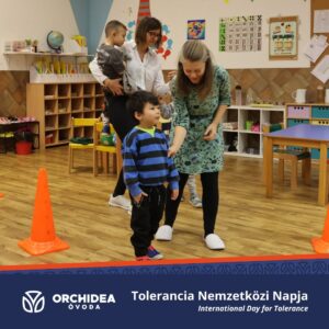 Egy kisfiú látszik munkatársunkkal, aki magyarázza neki a feladatot, mögöttük egy óvónéni is egy másik kisgyerekkel