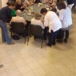 A februári szabadidős klub résztvevői az összetolt asztalok körül ülve fűzik a gyöngyöt, mellettük állnak segítőik, egy jeltolmács is jelel éppen