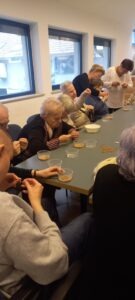 Szintén az asztalnál ülnek a tagok, mellettük a segítők, előttük átlátszó tálkában arany színű gyöngyök