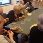 Szintén az asztalnál ülnek a tagok, mellettük a segítők, előttük átlátszó tálkában arany színű gyöngyök