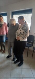 Az oktató közvetlenül az egyik résztvevő mellett áll, vállát átkarolva mutatja neki a csípőbillentés mozdulatát,saját testén is érzékeltetve,hogy kell pontosan végezni a gyakorlatot