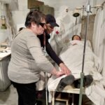 Az egyik résztvevő és a segítője megérinti az ágyban fekvő bábut, amely előtt egy infúziós állvány látható