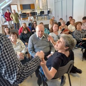 A résztvevők az Egyesületi nagy teremben ülnek egymás mellett, sorokban elrendezett székeken, jeltolmács is látszik, ahogy jelel és az érkezők is jönnek még lefelé a lépcsőn
