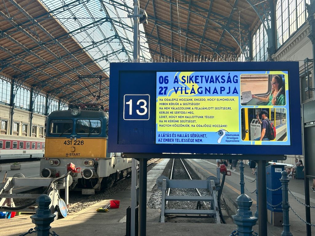 Pályaudvaron látszik egy vonatszerelvény és a kijelző, amin a siketvakság világnapi plakátunk jelenik meg