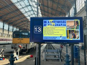 Pályaudvaron látszik egy vonatszerelvény és a kijelző, amin a siketvakság világnapi plakátunk jelenik meg