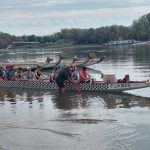 A sárkányhajó oldalról, benne a résztvevők, a túravezető az evezést tanítja a vízben állva, háttérben még több sárkányhajó