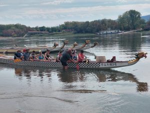 A sárkányhajó oldalról, benne a résztvevők, a túravezető az evezést tanítja a vízben állva, háttérben még több sárkányhajó