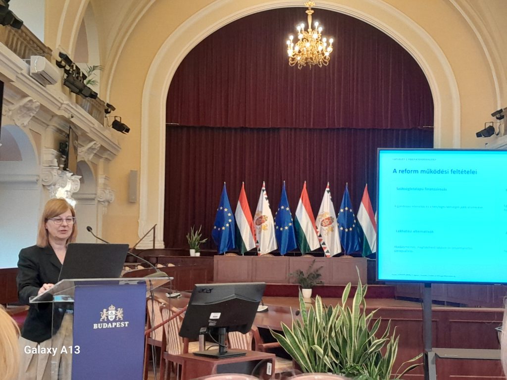 Bódi Éva előadás közben. Mellette a magyar, a Budapest városi és az EU-s zászló látszik. Egy kivetített kép is, melynek címsorában majd az alatt ez olvasható: A reform működési feltételei, a szükségletalapú finanszírozás